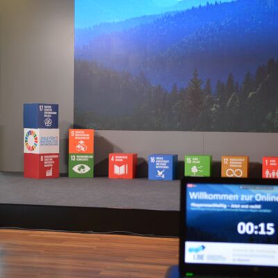 Bayerische Nachhaltigkeitstagung 2020: Bühne mit Hintergrundbild und SDG-Würfeln, im Vordergrund ist ein Bildschirm mit einem Countdown bis zum Start der Online-Tagung zu sehen