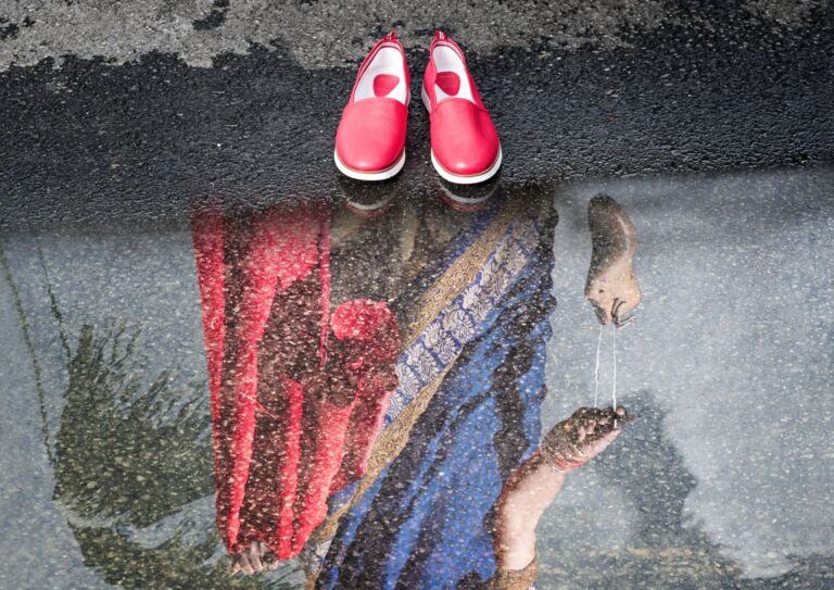 Ein Paar rote Schuhe steht vor einer Pfütze. In der Pfütze ist das Spiegelbild einer Frau in einem Sari-ähnlichen Gewand zu sehen, die einen Schuh in der Hand hält